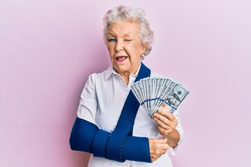 Senior grey-haired woman wearing arm on sling holding usa dollars banknotes winking looking at the camera with sexy expression, cheerful and happy face.