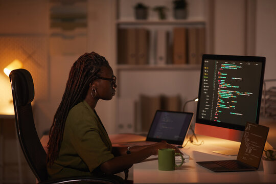 Side view portrait of contemporary African-American woman writing code and looking at computer screen while working in dim office, copy space