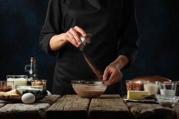 The chef in black apron pours cocoa into glass bowl for preparing dough. Backstage of cooking waffle on rustic wooden table with ingredients on dark blue background. Frozen motion. Traditional recipe.