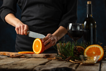 Chef cuts with knife juicy orange on wooden chopped board for preparing mulled wine on rustic wooden table with festive composition background. Backstage of cooking hot drink with fragrant spices.