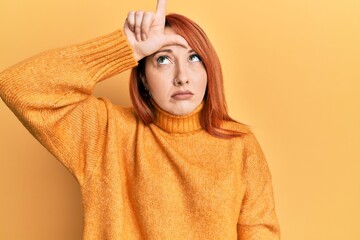 Beautiful redhead woman wearing casual winter sweater over yellow background making fun of people with fingers on forehead doing loser gesture mocking and insulting.