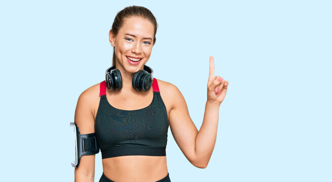 Beautiful blonde woman wearing gym clothes and using headphones pointing finger up with successful idea. exited and happy. number one.