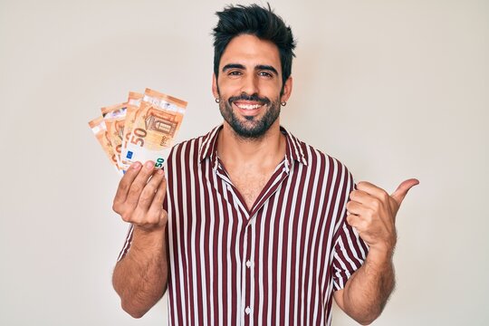 Handsome hispanic man with beard holding euro banknotes pointing thumb up to the side smiling happy with open mouth