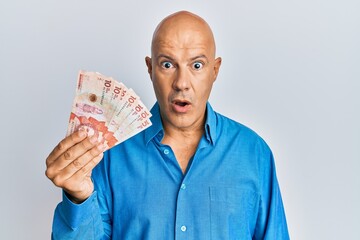 Middle age bald man holding 10 colombian pesos banknotes scared and amazed with open mouth for surprise, disbelief face