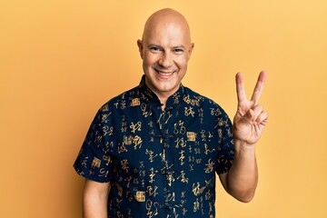 Middle age bald man wearing casual clothes smiling with happy face winking at the camera doing victory sign. number two.