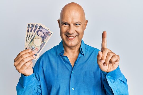 Middle age bald man holding 5000 japanese yen banknotes smiling with an idea or question pointing finger with happy face, number one