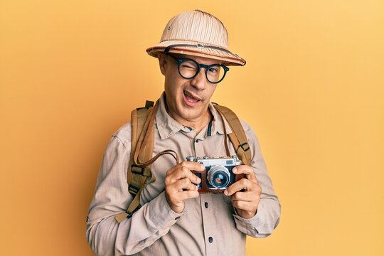 Middle age bald man wearing explorer hat and vintage camera winking looking at the camera with sexy expression, cheerful and happy face.