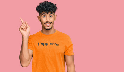 Young arab man wearing tshirt with happiness word message with a big smile on face, pointing with hand finger to the side looking at the camera.
