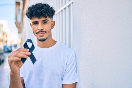Young arab man with serious expression holding mouring black ribbon leaning on the wall.