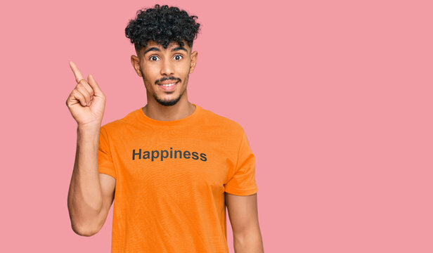 Young arab man wearing tshirt with happiness word message with a big smile on face, pointing with hand finger to the side looking at the camera.