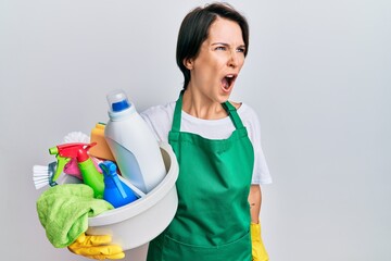 Young brunette woman with short hair wearing apron holding cleaning products angry and mad screaming frustrated and furious, shouting with anger. rage and aggressive concept.