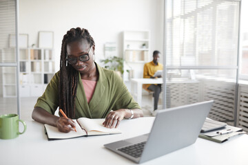 Fototapeta na wymiar Front view portrait of contemporary African-American woman writing in planner while working at desk in white office interior, copy space