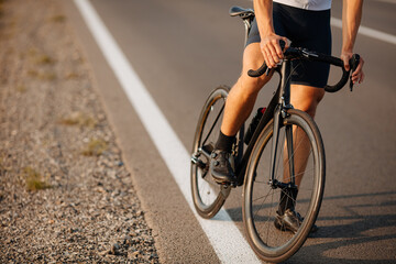 Fototapeta na wymiar Young sportsman in activewear and sneakers sitting on black bike on paved road. Close up of strong male legs. Regular training on fresh air.