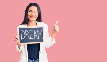 Young beautiful latin girl holding blackboard with dream word smiling happy and positive, thumb up doing excellent and approval sign