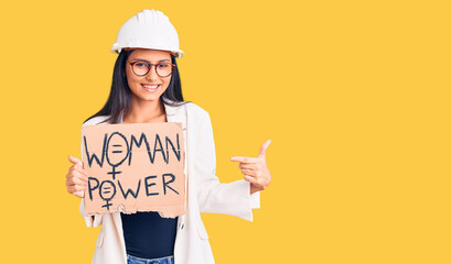 Young beautiful latin girl wearing architect hardhat holding woman power banner smiling happy...