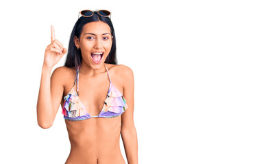 Obraz premium Young beautiful latin girl wearing bikini and sunglasses pointing finger up with successful idea. exited and happy. number one.
