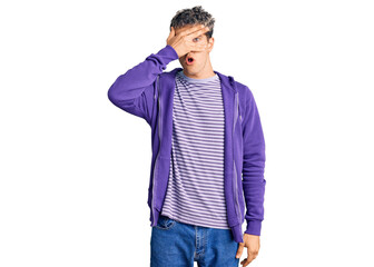 Fototapeta premium Young handsome man wearing casual purple sweatshirt peeking in shock covering face and eyes with hand, looking through fingers with embarrassed expression.
