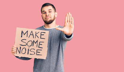 Young handsome man holding make some noise banner with open hand doing stop sign with serious and...