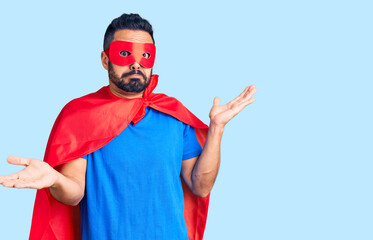 Young hispanic man wearing super hero costume clueless and confused expression with arms and hands...