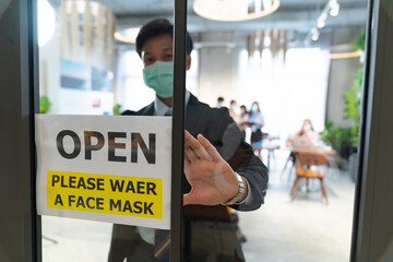 A businessman wearing a surgical mask puts an open sign "Open" at the front door, reopening business to adapt to the new normal in preventing the spread of the COVID-19 coronavirus.