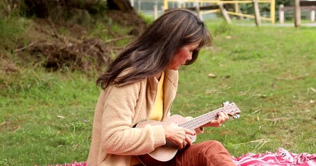 Relaxing attractive long-haired woman wearing casual wear and sitting on a blanket at a park learning to play the ukulele she has on her knee. Medium profile shot.