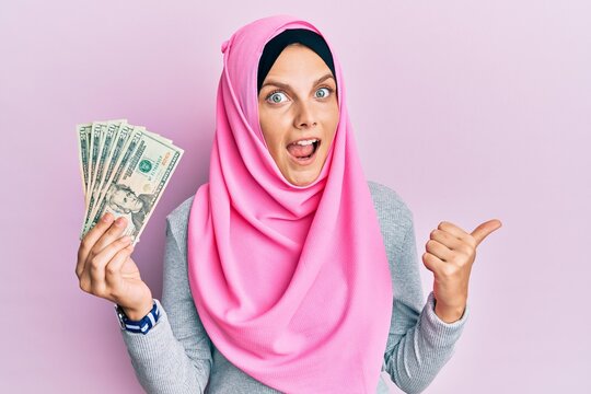 Young caucasian woman wearing islamic hijab holding 20 dollars banknotes pointing thumb up to the side smiling happy with open mouth