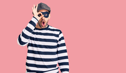 Young handsome man wearing burglar mask doing ok gesture shocked with surprised face, eye looking...