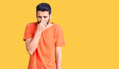 Young handsome man with beard wearing casual t-shirt smelling something stinky and disgusting,...