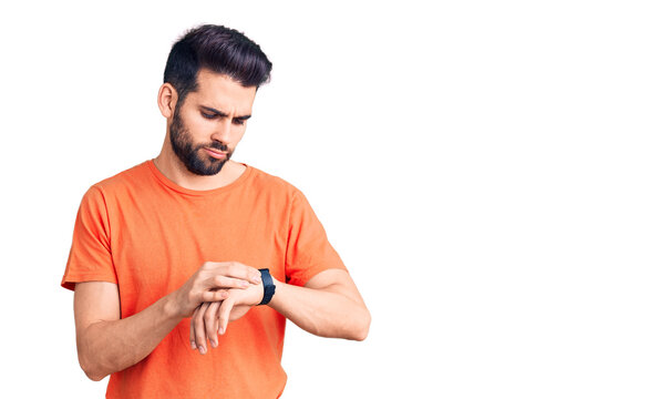 Young handsome man with beard wearing casual t-shirt checking the time on wrist watch, relaxed and confident