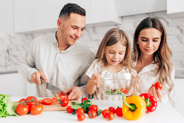 Happy family of little girl and her parents having fun and cooking salad in kitchen at home.