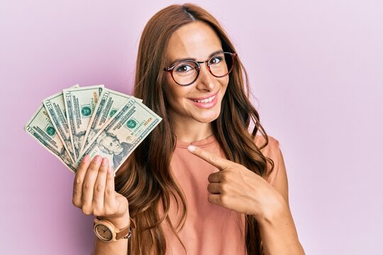 Young brunette woman holding dollars smiling happy pointing with hand and finger