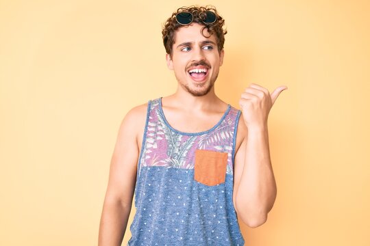 Young caucasian man with curly hair wearing casual style with sleeveless shirt pointing thumb up to the side smiling happy with open mouth