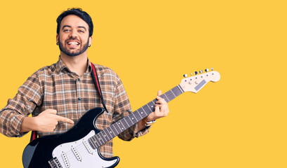 Young hispanic man playing electric guitar smiling happy pointing with hand and finger