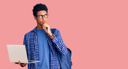 Young african american man holding student backpack using laptop serious face thinking about...
