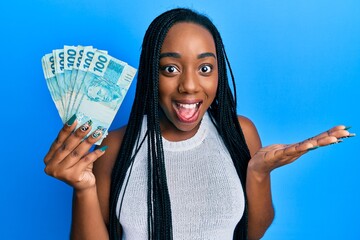 Young african american woman holding 100 brazilian real banknotes celebrating achievement with...