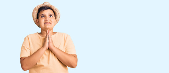 Little boy kid wearing summer hat and hawaiian swimsuit begging and praying with hands together with hope expression on face very emotional and worried. begging.