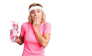 Young blonde woman wearing sportswear holding water bottle covering mouth with hand, shocked and...