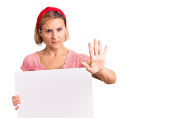 Young blonde woman holding blank empty banner with open hand doing stop sign with serious and...