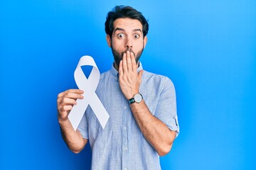 Young hispanic man holding blue ribbon covering mouth with hand, shocked and afraid for mistake....