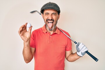 Middle age hispanic man holding golf club and ball sticking tongue out happy with funny expression.