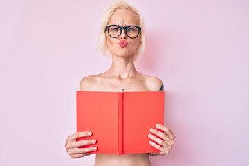 Young blonde woman with tattoo standing shirtless reading book making fish face with mouth and squinting eyes, crazy and comical.
