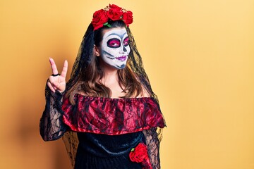 Young woman wearing day of the dead costume over yellow smiling with happy face winking at the camera doing victory sign. number two.