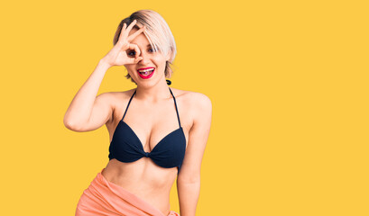 Naklejka premium Young beautiful blonde woman wearing bikini smiling happy doing ok sign with hand on eye looking through fingers