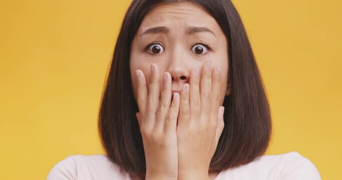 Young shocked surprised asian woman closing mouth in amazement, looking worried and stunned, orange studio background