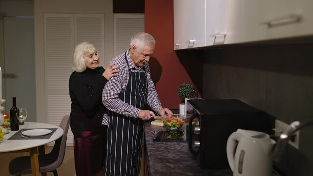 Side view of happy mature couple in love making dinner. Elderly woman hugging from back her husband cooking meal standing near cutting board in stylish kitchen at home. Senior grandmother, grandfather