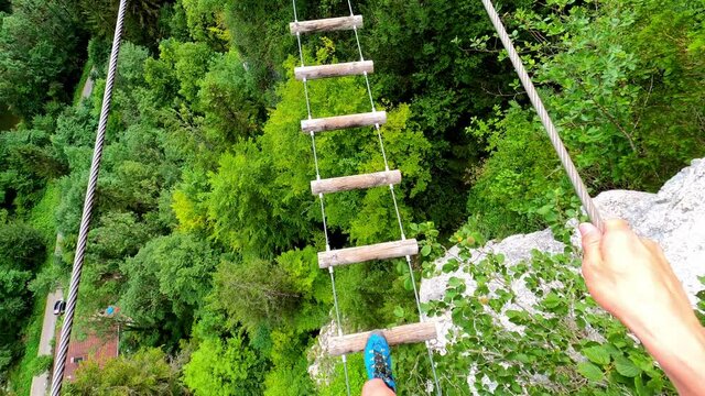 Am man walking on a suspension bridge made out of pieces of wood. Underneath is a forest. Its the via ferrata "ladders of death" in France.