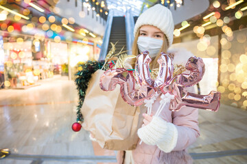 Young woman in medical mask shopping for Christmas in mall. Female holding golden balloons in form of numbers 2021. Xmas holidays in new Covid-19 reality. Selective focus