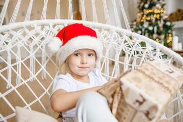 A four-year-old girl in a Santa hat sits in a wicker chair against the background of a Christmas tree and unwraps a gift