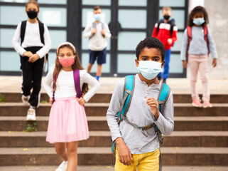 Portrait of schoolboy in medical mask standing near school, kids on background ..
