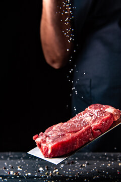 the cook holds in his hand a butcher's knife with a steak and sprinkles it with salt. makes the final touch to the dish before cooking. Vertical photo. With free space for text, ad labels and logos